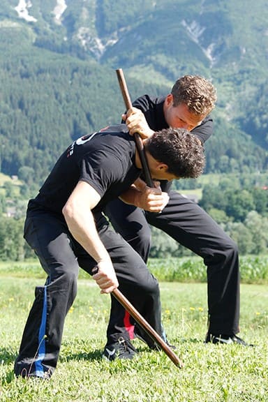 Partnerübung mit einem Stock zur Demonstration einer Hebel- oder Würgetechnik beim Waffentraining im Freien der Akademie für Selbstverteidigung, Kampfkünste und Kampfsport in Graz.